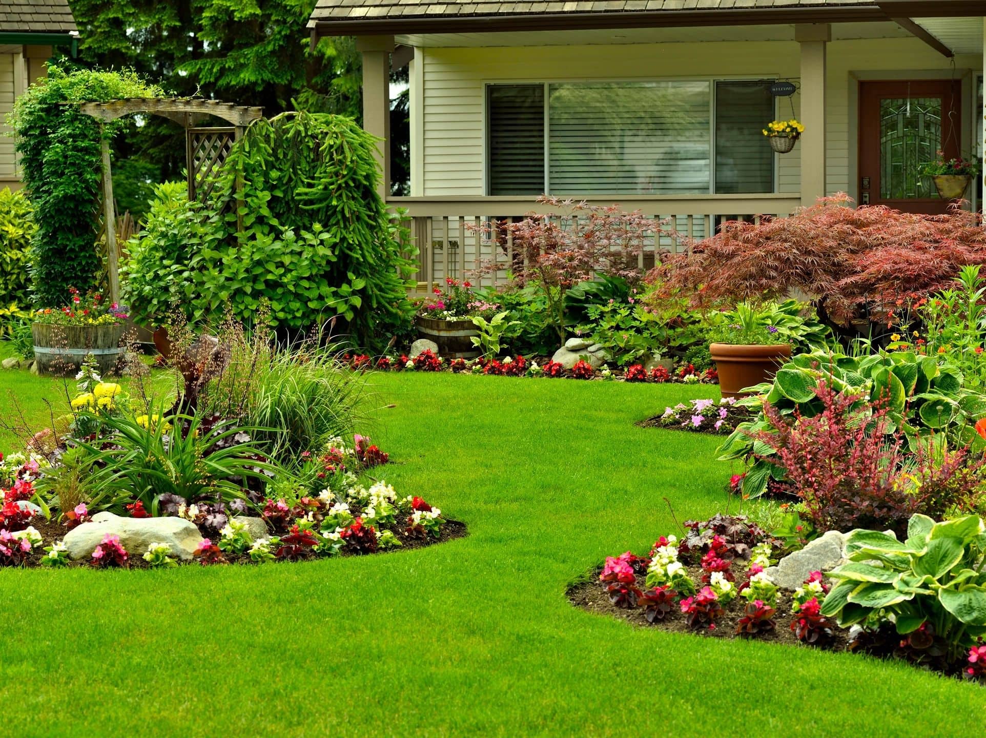 A lush green lawn with vibrant flower beds, various shrubs, and potted plants surrounds the front of a house with a porch, large window, and wooden door.