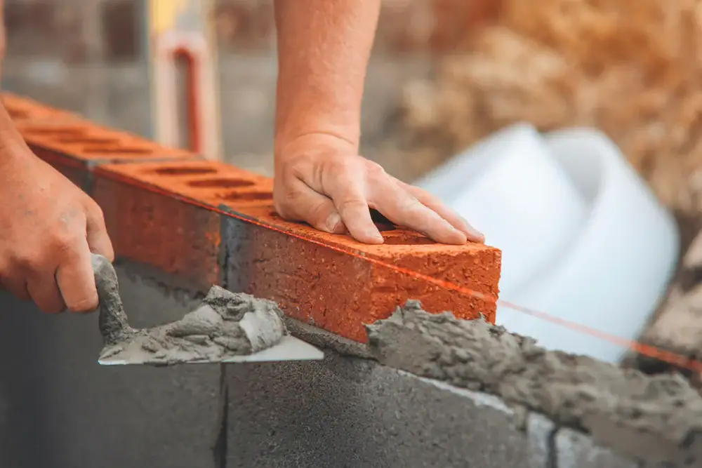 A close-up of a person laying red bricks, using a trowel to spread cement mortar on a gray concrete wall, with hands positioning the bricks carefully.
