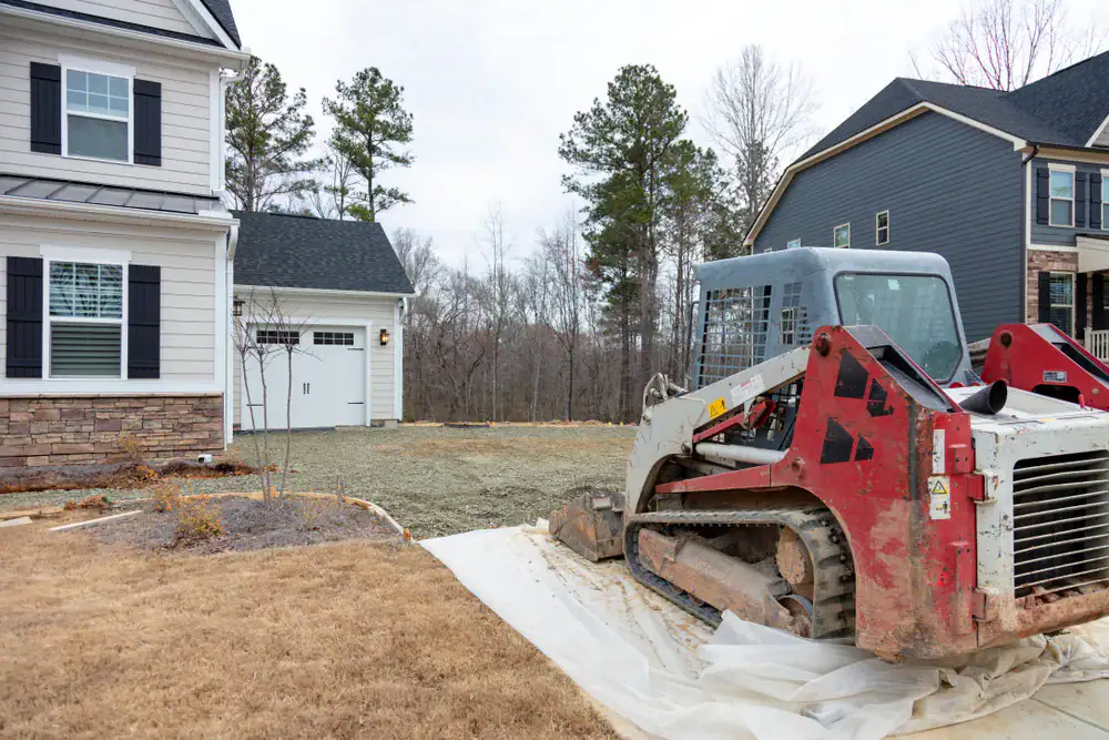A small bulldozer sits on a gravel driveway under construction between two suburban houses, with trees and overcast sky in the background.