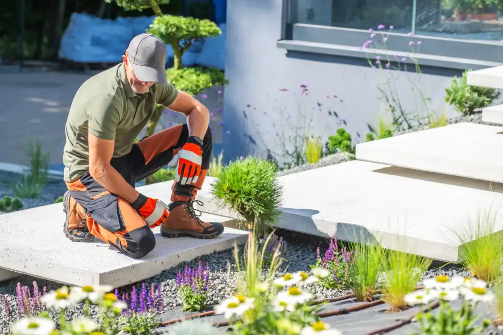 A gardener wearing gloves, a cap, and protective kneepads tends to plants and flowers beside modern concrete steps in a landscaped garden.