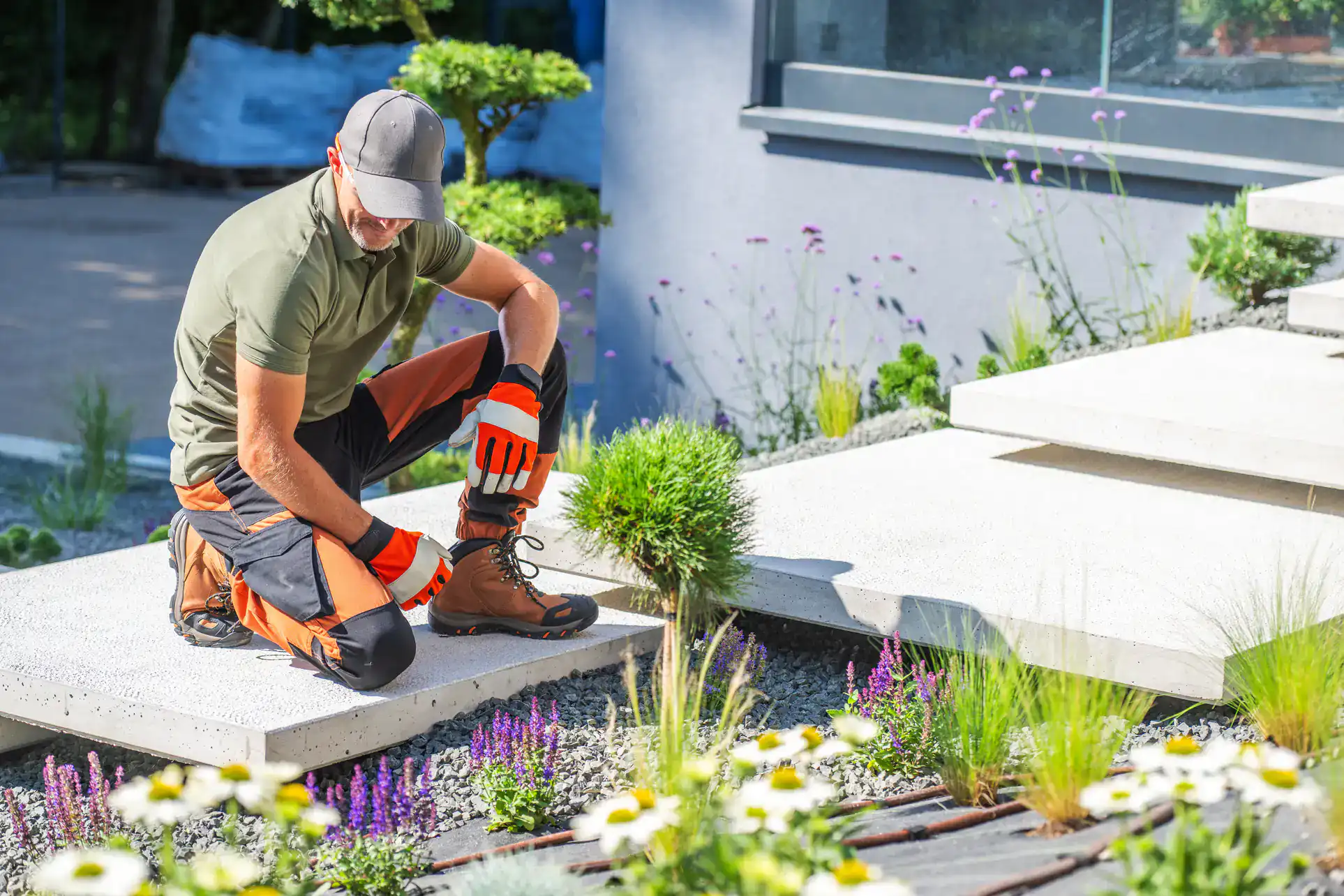 A gardener wearing gloves, a cap, and protective kneepads tends to plants and flowers beside modern concrete steps in a landscaped garden.