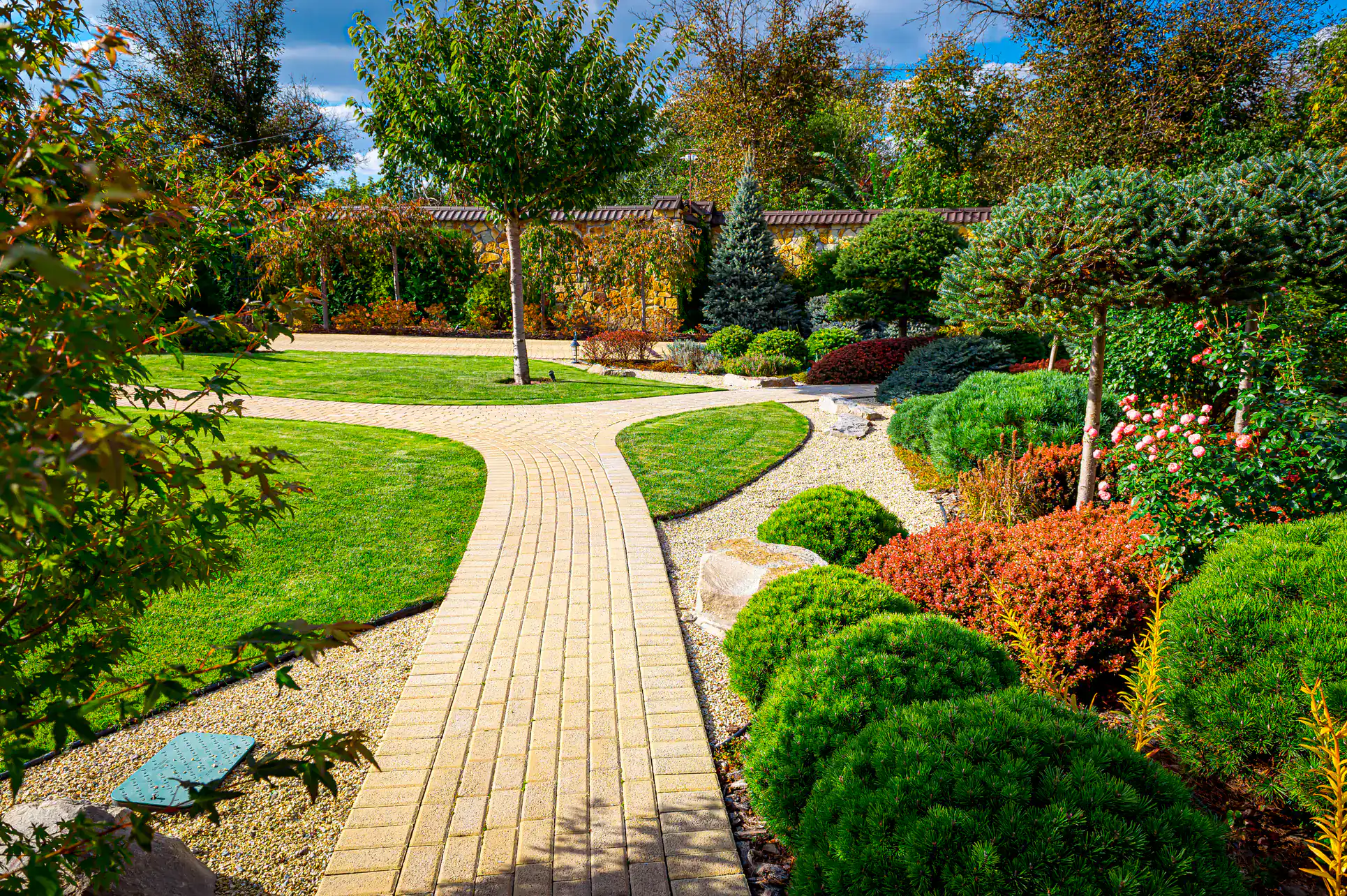 A beautifully landscaped garden featuring a winding brick pathway, neatly trimmed lawns, colorful shrubs, vibrant flowers, small trees, and a wooden fence in the background under a blue sky.