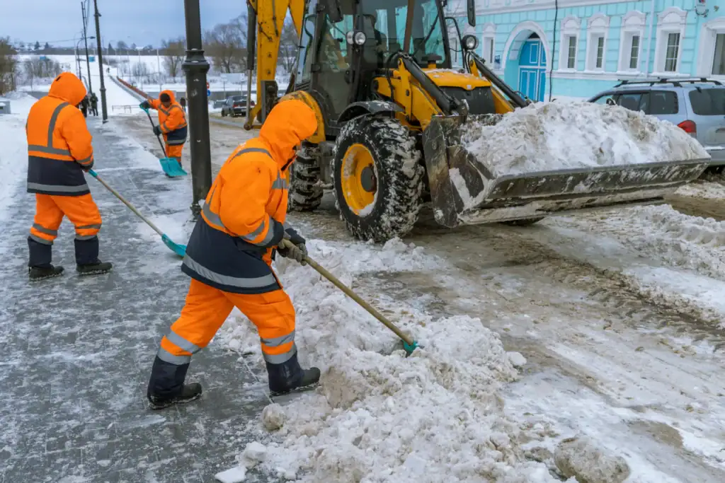 Three workers in bright orange winter uniforms shovel snow from a road, while a backhoe loader collects snow in its front bucket. Snow-covered ground and parked cars are visible in the background.