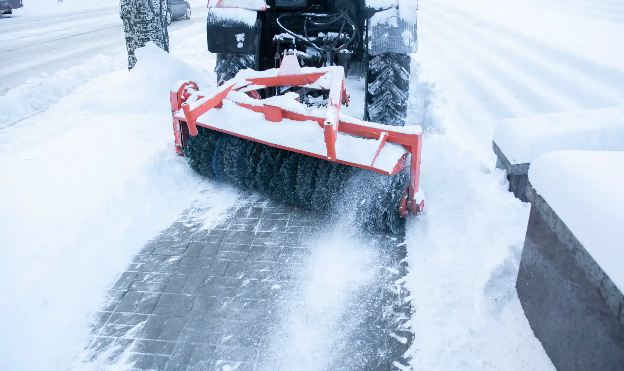 A snow removal machine with a rotating brush attachment clears snow from a paved sidewalk on a cold, snowy day.