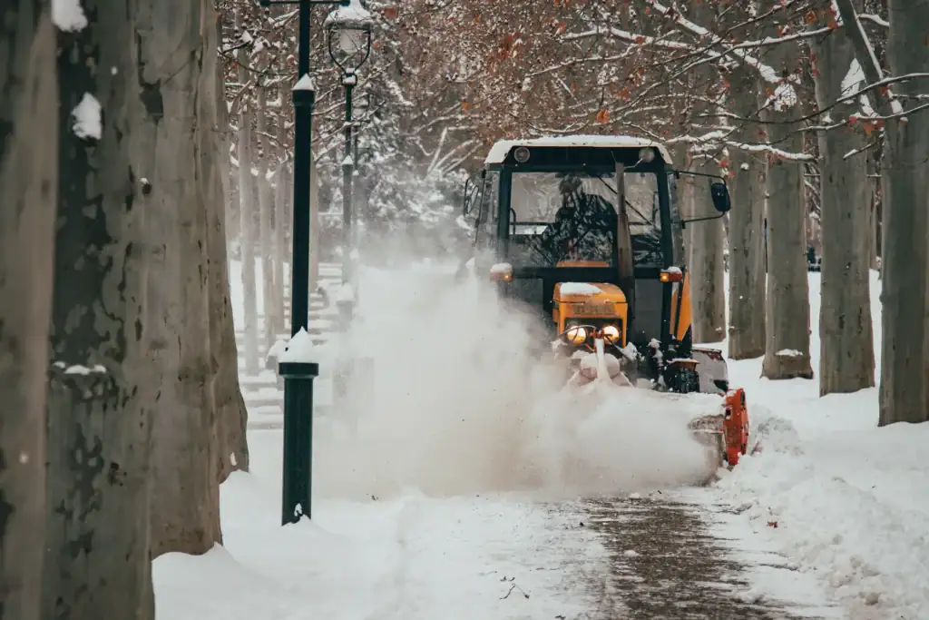 A snowplow clears a snow-covered path in a park lined with bare trees on a winter day, with snow blowing up around the vehicle as it moves forward.