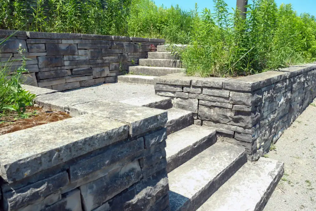 Stone steps and walls with stacked flat rocks lead up a terraced area surrounded by tall green plants and grass, under bright sunlight and clear blue sky.