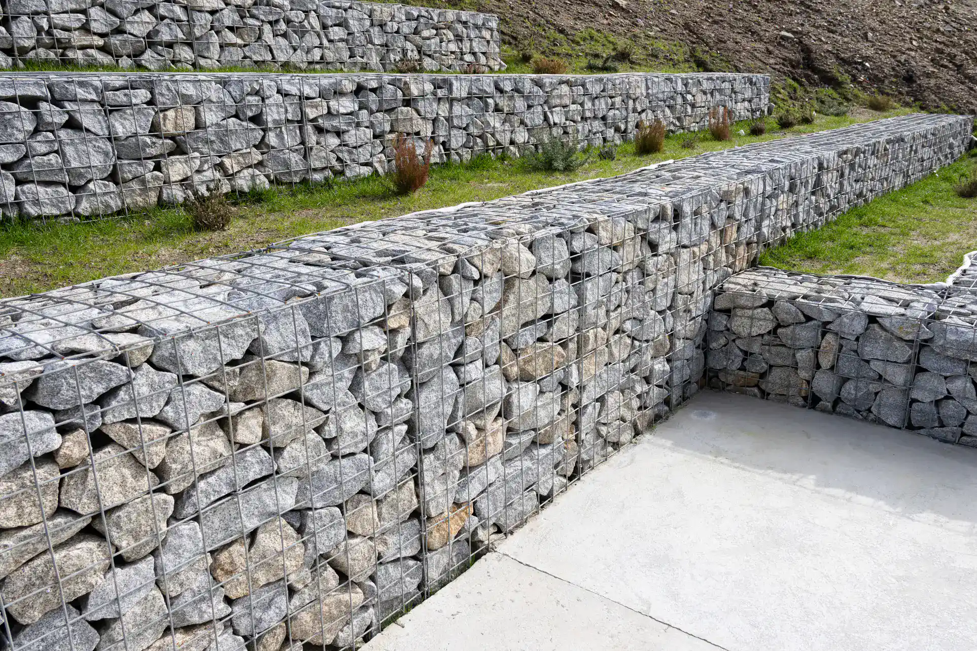 Stone-filled wire gabion walls are arranged in terraces on a sloped outdoor landscape with patches of grass between them and a concrete surface in the foreground.