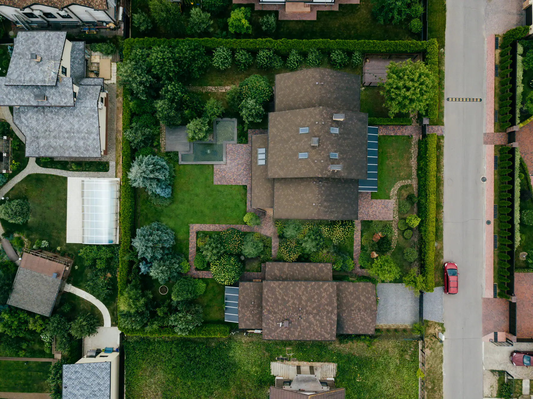 Aerial view of a suburban neighborhood with houses, green lawns, trees, and gardens separated by hedges. A red car is parked on the road next to a property with brown roofs and brick paths.