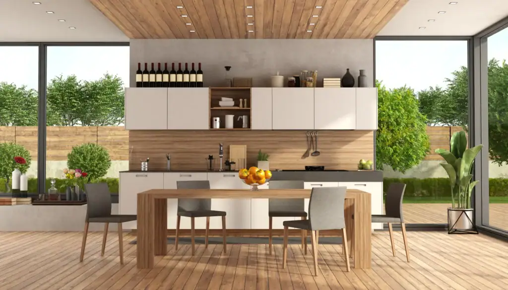 Modern kitchen with wooden floors and ceiling, white cabinets, a wooden dining table with four gray chairs, large windows showing a green garden, and bottles of wine on the top shelf. A bowl of fruit is on the table.