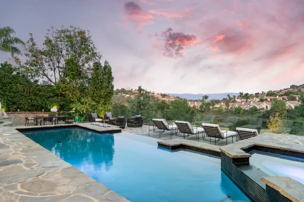 A luxurious backyard with a stone patio, modern pool, hot tub, lounge chairs, and lush greenery, overlooking a scenic hillside neighborhood at sunset with dramatic clouds in the sky.